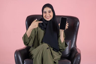 عرض تطبيقات الجوال، الاشارة باصبع السبابة الى الجوال، النظر الى الكاميرا بايماءات السعادة والسرور، عرض شاشة سوداء فارغة، بورتريه لامرأة عربية خليجية اماراتية محجبة ترتدي العباءة تجلس على كرسي تمسك بيدها الهاتف المحمول، استخدام جهاز تقني حديث ومتطور، خلفية وردية