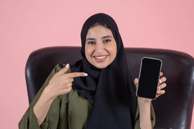 Pointing with the index finger at the mobile phone, looking at the camera with gestures of happiness and joy, displaying a blank black screen, showcasing mobile applications, a close-up portrait of a veiled Emirati Gulf Arab woman wearing an abaya sitting on a chair holding a mobile phone in her hand, using a modern and advanced technological device, pink background.