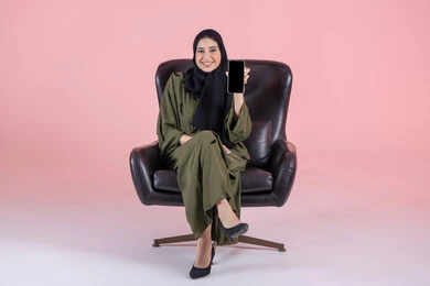 النظر الى الكاميرا بايماءات السعادة والسرور، عرض شاشة سوداء فارغة، عرض تطبيقات الجوال، بورتريه لامرأة عربية خليجية اماراتية محجبة ترتدي العباءة تجلس على كرسي تمسك بيدها الهاتف المحمول، استخدام جهاز تقني حديث ومتطور، خلفية وردية