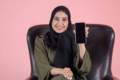 A blank black screen is displayed, looking at the camera with gestures of happiness and joy, showcasing mobile applications, a portrait of an Emirati Gulf Arab woman wearing a hijab and an abaya sitting on a chair holding a mobile phone in her hand, using a modern and advanced technological device, with a pink background.