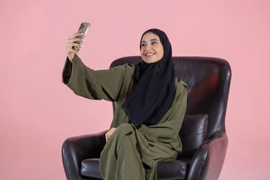Using a modern technical device, documenting happy moments, taking selfies with a mobile phone, a portrait of an Emirati Gulf Arab woman wearing a hijab and abaya sitting on a chair holding a mobile phone with expressions of joy and happiness, pink background.