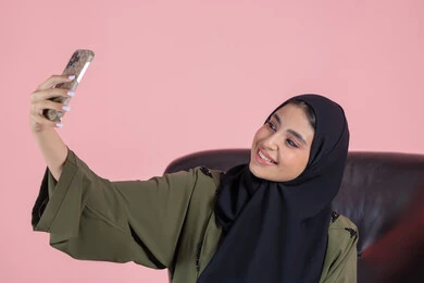 Documenting happy moments, taking selfies using a mobile phone, a close-up portrait of an Emirati Gulf Arab woman wearing a hijab and abaya sitting on a chair holding a mobile phone with expressions of joy and happiness, using modern technology, pink background.