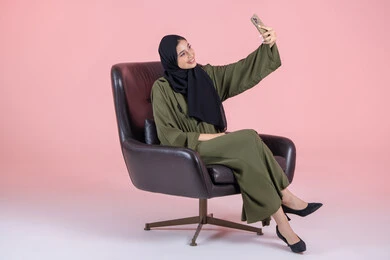 Preserving special memories through photography, taking selfies using a mobile phone, a portrait of an Emirati Gulf Arab woman wearing a hijab and abaya sitting on a chair holding a mobile phone in her hand with expressions of joy and happiness, using modern technology, pink background.