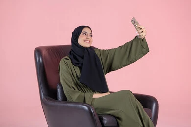 حفظ الذكريات الجميلة بعدسة الكاميرا، التقاط صور سيلفي باستخدام الهاتف المحمول، بورتريه لإمرأة عربية خليجية امارتية ترتدي الحجاب والعباءة  تجلس على كرسي تحمل بيدها الجوال بتعابير السرور والسعادة، استخدام جهاز تقني حديث، خلفية وردية