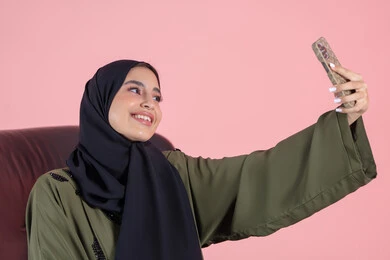 تسجيل اللحظات المميزة، التقاط صور سيلفي باستخدام الهاتف المحمول، بورتريه مقرب لإمرأة عربية خليجية امارتية ترتدي الحجاب والعباءة  تجلس على كرسي تحمل بيدها الجوال بتعابير السرور والسعادة، استخدام جهاز تقني حديث، خلفية وردية