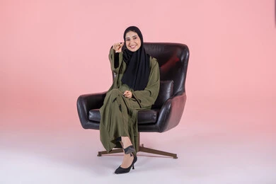 Pointing to a specific location, expressions of happiness and joy, identifying an element, a portrait of an Emirati Gulf Arab woman wearing a hijab and abaya sitting on a chair pointing with her hands to something, pink background.