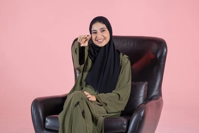 Accurately identifying the desired location through guidance, expressions of happiness and joy, identifying an element, a portrait of an Arab Gulf Emirati woman wearing a hijab and abaya sitting on a chair pointing with her hands to something, pink background.