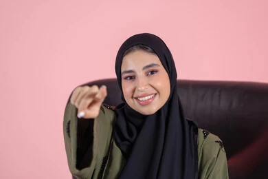 توضيح موقع معين، تعابير السعادة و السرور، تحديد عنصر ما، بورتريه مقرب لامرأة عربية خليجية اماراتية ترتدي الحجاب والعباءة تجلس على كرسي تشير بيديها الى شيء ما, خلفية وردية