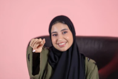 تعابير السعادة و السرور، الإشارة إلى مكان محدد، تحديد عنصر ما، بورتريه مقرب لامرأة عربية خليجية اماراتية ترتدي الحجاب والعباءة تجلس على كرسي تشير بيديها الى شيء ما, خلفية وردية