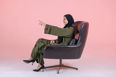 الإشارة إلى مكان محدد،  تعابير السعادة و السرور، تحديد عنصر ما، بورتريه من الجانب لامرأة عربية خليجية اماراتية ترتدي الحجاب والعباءة تجلس على كرسي تشير بيديها الى شيء ما, خلفية وردية