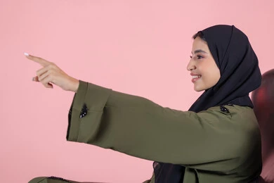 Directing attention to a specific point, expressions of happiness and joy, pointing to an element, a side portrait of an Arab Gulf Emirati woman wearing a hijab and abaya sitting on a chair pointing with her hands to something, pink background.