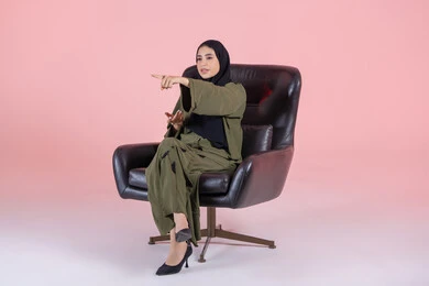 Identifying the location of a specific object, pointing to an element, clarifying and indicating a specific place, a portrait of an Arab Gulf Emirati woman wearing a hijab and abaya sitting on a chair pointing with her hands to something, pink background.