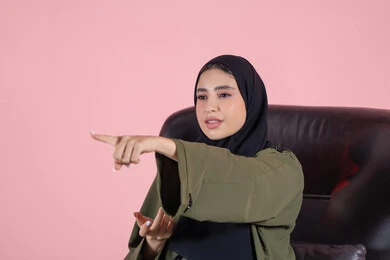 التوضيح والدلالة لمكان محدد, الإشارة إلى عنصر ما, بورتريه مقرب لامرأة عربية خليجية اماراتية ترتدي الحجاب والعباءة تجلس على كرسي تشير بيديها الى شيء ما، تحديد موقع شيء معين, خلفية وردية