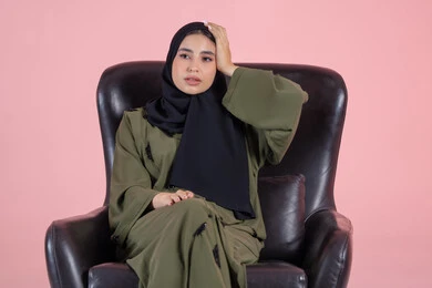 The attempt to remember something, an immediate recall of something necessary, gestures and facial and hand expressions indicating forgetfulness, disappointment, and regret, a close-up portrait of an Emirati Gulf Arab woman wearing a hijab and abaya sitting on a chair with her hand on her head, pink background.