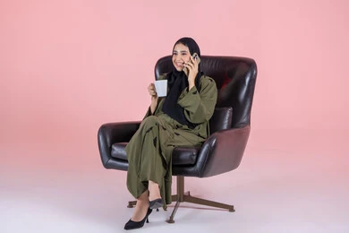 الحديث مع الأصدقاء وتبادل الأحاديث الممتعة، الإجابة والرد على الاتصالات بإستخدام الهاتف المحمول، تذوق المشروب المفضل، بورتريه لامرأة عربية خليجية اماراتية ترتدي الحجاب والعباءة تجلس على كرسي تقوم بإجراء مكالمة صوتية عبر الجوال، خلفية وردية