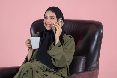التفاعل الاجتماعي وتبادل الأحاديث الممتعة، الإجابة والرد على الاتصالات بإستخدام الهاتف المحمول، تذوق المشروب المفضل، بورتريه لامرأة عربية خليجية اماراتية ترتدي الحجاب والعباءة تجلس على كرسي تقوم بإجراء مكالمة صوتية عبر الجوال، خلفية وردية