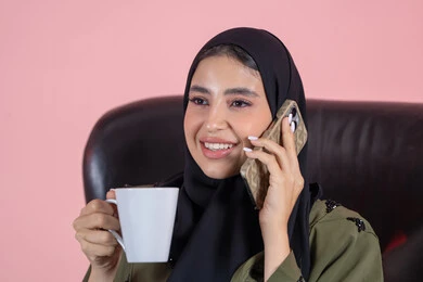 قضاء وقت جميل في التحدث ومشاركة الأخبار، الإجابة والرد على الاتصالات بإستخدام الهاتف المحمول، تذوق المشروب المفضل، بورتريه مقرب لامرأة عربية خليجية اماراتية ترتدي الحجاب والعباءة تجلس على كرسي تقوم بإجراء مكالمة صوتية عبر الجوال، خلفية وردية