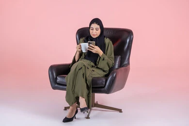 Spending enjoyable time, browsing websites and social media, online shopping, using electronic programs and applications, tasting the favorite drink, a portrait of an Emirati Gulf Arab woman wearing a hijab and abaya sitting on a chair and browsing a mobile phone, pink background.