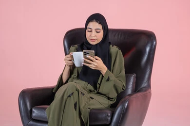 تصفح المواقع الالكترونية والتواصل الاجتماعي، التسوق الالكتروني عبر الانترنت، استخدام البرامج والتطبيقات الالكترونية، قضاء وقت ممتع،  تذوق المشروب المفضل, بورتريه لإمرأة عربية خليجية امارتية ترتدي الحجاب والعباءة تجلس على كرسي وتقوم بتصفح الهاتف المحمول، خلفية وردية