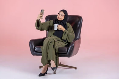 Taking selfies with a mobile phone, documenting beautiful moments, tasting a favorite drink, a portrait of a Gulf Arab Emirati woman wearing a hijab and abaya sitting on a chair holding a mobile phone with expressions of joy and happiness, using modern technology, pink background.