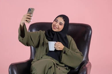 Documenting beautiful moments, tasting the favorite drink, taking selfies using a mobile phone, a portrait of a Gulf Arab Emirati woman wearing a hijab and abaya sitting on a chair holding a mobile phone with expressions of joy and happiness, using modern technology, pink background.