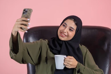 Taste the favorite drink, document beautiful moments, take selfies using a mobile phone, a close-up portrait of an Emirati Gulf Arab woman wearing a hijab and abaya sitting on a chair holding a mobile phone with expressions of joy and happiness, using modern technology, pink background.