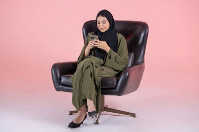 الدردشة مع الأصدقاء والأقارب عبر الجوال، استخدام جهاز تقني حديث ومتطور، تصفح مواقع التواصل الاجتماعي، بورتريه لشابة عربية خليجية اماراتية ترتدي الحجاب و العباءة تجلس على كرسي تمسك بيدها الهاتف المحمول، خلفية وردية