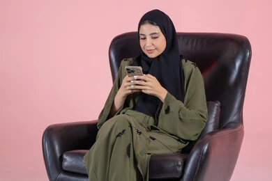 Using a modern and advanced technical device, browsing social media sites, chatting with friends and relatives via mobile, a portrait of a young Arab Gulf Emirati woman wearing a hijab and abaya sitting on a chair holding a mobile phone in her hand, pink background.
