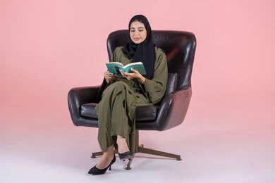 Spending useful time in self-development and knowledge enhancement, the concept of education and scientific research, the hobby of reading and exploration, a portrait of a young Arab Gulf Emirati woman wearing a hijab and abaya sitting on a chair reading a book, with a pink background.