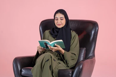 التعلم واكتساب المعلومات الجديدة أثناء وقت الفراغ، مفهوم التثقيف والبحث العلمي، هواية القراءة والاستكشاف, بورتريه لشابة عربية خليجية اماراتية ترتدي الحجاب و العباءة تجلس على كرسي تقوم بقراة كتاب، خلفية وردية