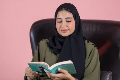تنمية الفكر والمعرفة خلال أوقات الفراغ، مفهوم التثقيف والبحث العلمي، هواية القراءة والاستكشاف, بورتريه مقرب لشابة عربية خليجية اماراتية ترتدي الحجاب و العباءة تجلس على كرسي تقوم بقراة كتاب، خلفية وردية