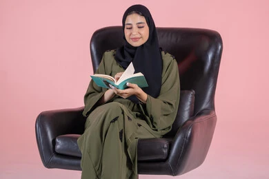 مفهوم التثقيف والبحث العلمي، هواية القراءة والاستكشاف، قضاء وقت الفراغ بالتعلم والثقافة, بورتريه لشابة عربية خليجية اماراتية ترتدي الحجاب و العباءة تجلس على كرسي تقوم بقراة كتاب، خلفية وردية