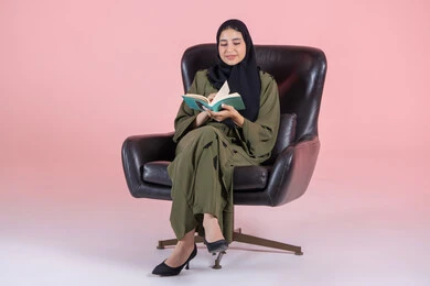 The hobby of reading and exploration, spending free time learning and cultural activities, the concept of education and scientific research, a portrait of a young Arab Gulf Emirati woman wearing a hijab and abaya sitting on a chair reading a book, with a pink background.