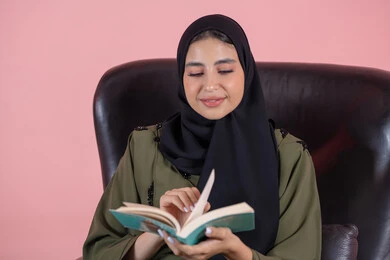 Spending free time on learning and culture, the hobby of reading and exploration, the concept of education and scientific research, a close-up portrait of a young Arab Gulf Emirati woman wearing a hijab and abaya sitting on a chair reading a book, with a pink background.