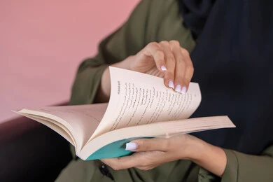 Interest in science and culture, developing and enhancing general knowledge, the hobby of reading and exploration, a close-up portrait of a young Arab Gulf Emirati woman holding a book in her hand, with a pink background.
