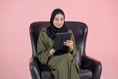 E-learning through the use of modern technologies, making video calls and live broadcasts, watching favorite movies and series online, tapping on the tablet screen, a portrait of a young Arab Gulf Emirati woman wearing a hijab and abaya sitting on a chair holding a mobile tablet, pink background.