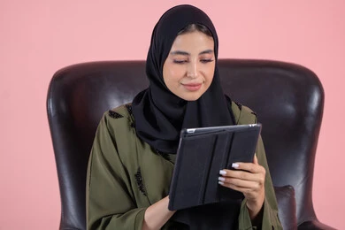 Making video calls and live broadcasts, watching favorite movies and series online, tapping on the tablet screen, a close-up portrait of a young Emirati Gulf Arab woman wearing a hijab and abaya sitting on a chair holding a portable tablet, e-learning through the use of modern technologies, pink background.