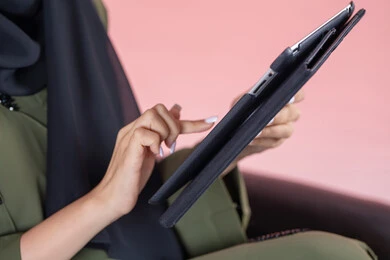 Using the touch feature, hand gestures by tapping fingers on the screen, using modern technology devices, a close-up side portrait of a young Arab Gulf Emirati woman holding a tablet, with a pink background. Using the touch feature, hand gestures by tapping fingers on the screen, using modern technology devices, a close-up side portrait of a young Arab Gulf Emirati woman holding a tablet, with a pink background.