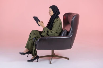 Watching favorite movies and series online, tapping on the tablet screen, a side portrait of a young Arab Gulf Emirati woman wearing a hijab and abaya sitting on a chair holding a portable tablet, making video calls and live broadcasts, e-learning through the use of modern technologies, pink background.