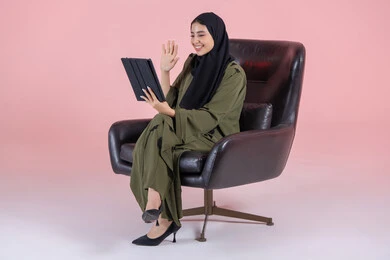 Communicating with friends, expressions of happiness and joy, raising a hand in gestures of peace and greeting, using advanced modern technology in professional life through a tablet, a side portrait of a young Emirati Gulf Arab woman wearing a hijab and abaya sitting on a chair working on a tablet, with a pink background.