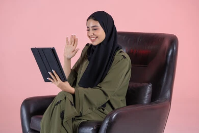 اجراء مكالمة فيديو عبر الإنترنت، تعابير السعادة و السرور، رفع كف الايد بايماءات السلام و التحيه، استخدام التقنية الحديثه المتطورة في الحياة العملية عبر الجهاز اللوحي، بورتريه من الجانب لشابة عربية خليجية اماراتية ترتدي الحجاب والعباءة تجلس على كرسي تقوم بالعمل على جهازالتابلت، خلفية وردية