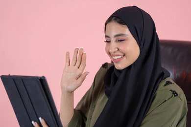 تعابير السعادة و السرور، رفع كف الايد بايماءات السلام و التحيه، استخدام التقنية الحديثه المتطورة في الحياة العملية عبر الجهاز اللوحي، بورتريه مقرب من الجانب لشابة عربية خليجية اماراتيةترتدي الحجاب والعباءة تجلس على كرسي تقوم بالعمل على جهازالتابلت، اجراء مكالمات فيديو عبر الإنترنت، خلفية وردية