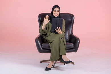 The use of advanced modern technology in professional life through a tablet, raising the hand in gestures of peace and greeting, expressions of happiness and joy, a portrait of a young Arab Gulf Emirati woman wearing a hijab and abaya sitting on a chair working on a tablet, making video calls over the internet, with a pink background.