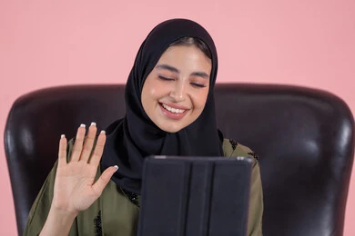رفع كف الايد بايماءات السلام و التحيه، استخدام التقنية الحديثه المتطورة في الحياة العملية عبر الجهاز اللوحي، تعابير السعادة و السرور، بورتريه مقرب لشابة عربية خليجية اماراتيةترتدي الحجاب والعباءة تجلس على كرسي تقوم بالعمل على جهازالتابلت، اجراء مكالمات فيديو عبر الإنترنت، خلفية وردية