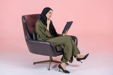 Shopping through technical devices, expressions and gestures of integration and thinking about something, a side portrait of a young Arab Gulf Emirati woman wearing a hijab and abaya sitting on a chair working on a tablet device, with a pink background.