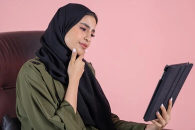 الدردشة والتواصل مع الأهل والأصدقاء، تعابير وايماءات الاندماج و التفكير فى شيء ما، بورتريه من الجانب لشابة عربية خليجية اماراتية ترتدي الحجاب والعباءة تجلس على كرسي تقوم بالعمل على جهازالتابلت،خلفية وردية