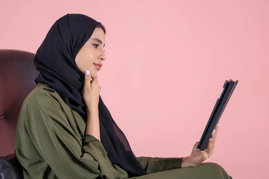 استخدام التقنية الحديثه المتطورة في الحياة العملية عبر الجهاز اللوحي، تعابير وايماءات الاندماج و التفكير فى شيء ما، بورتريه من الجانب لشابة عربية خليجية اماراتية ترتدي الحجاب والعباءة تجلس على كرسي تقوم بالعمل على جهازالتابلت،خلفية وردية