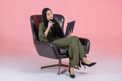 انجاز مهام العمل عبر التابلت، اللعب على الجهاز المحمول، بورتريه لإمرأه عربيه خليجيه اماراتية تجلس على كرسي مريح, إدارة المشاريع عن بعد, إجراء مكالمات فيديو عبر الإنترنت, مشاهدة فلم, تسليه وترفيه, قضاء وقت ممتع، خلفية وردية