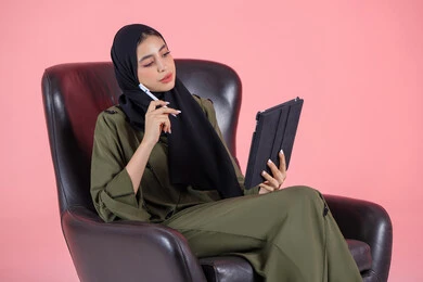 اللعب على الجهاز المحمول، بورتريه لإمرأه عربيه خليجيه اماراتية تجلس على كرسي مريح, انجاز مهام العمل عبر التابلت, إدارة المشاريع عن بعد, إجراء مكالمات فيديو عبر الإنترنت, مشاهدة فلم, تسليه وترفيه, قضاء وقت ممتع، خلفية وردية