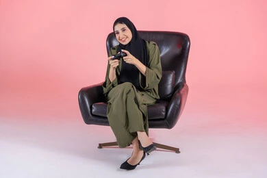 Enjoying digital entertainment during the weekend, gestures of immersion and interaction while playing, using modern and advanced electronic technologies, a portrait of an Arab Gulf Emirati woman wearing an abaya and hijab sitting on a chair holding a game controller, with a pink background.